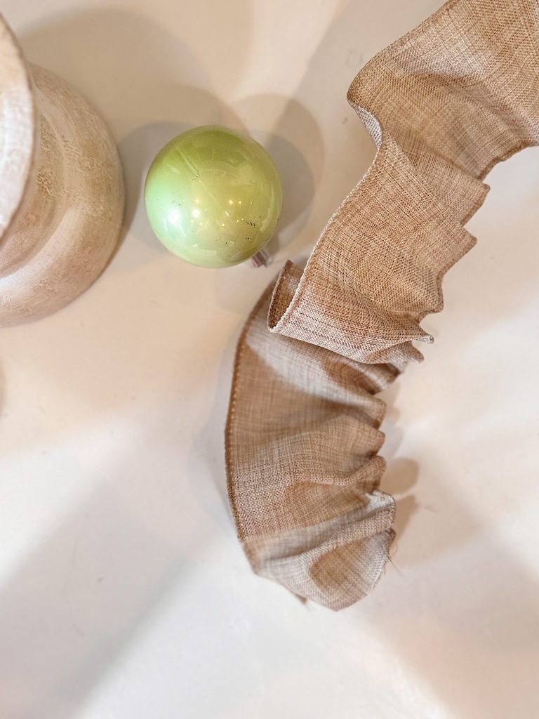 A green ornament, a wooden object, and a ruffled strip of burlap fabric are arranged on a white surface.