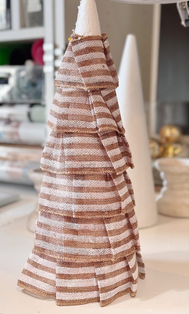 A cone-shaped structure covered in layered, striped brown and white fabric is displayed on a table, with another plain white cone and craft materials in the background.