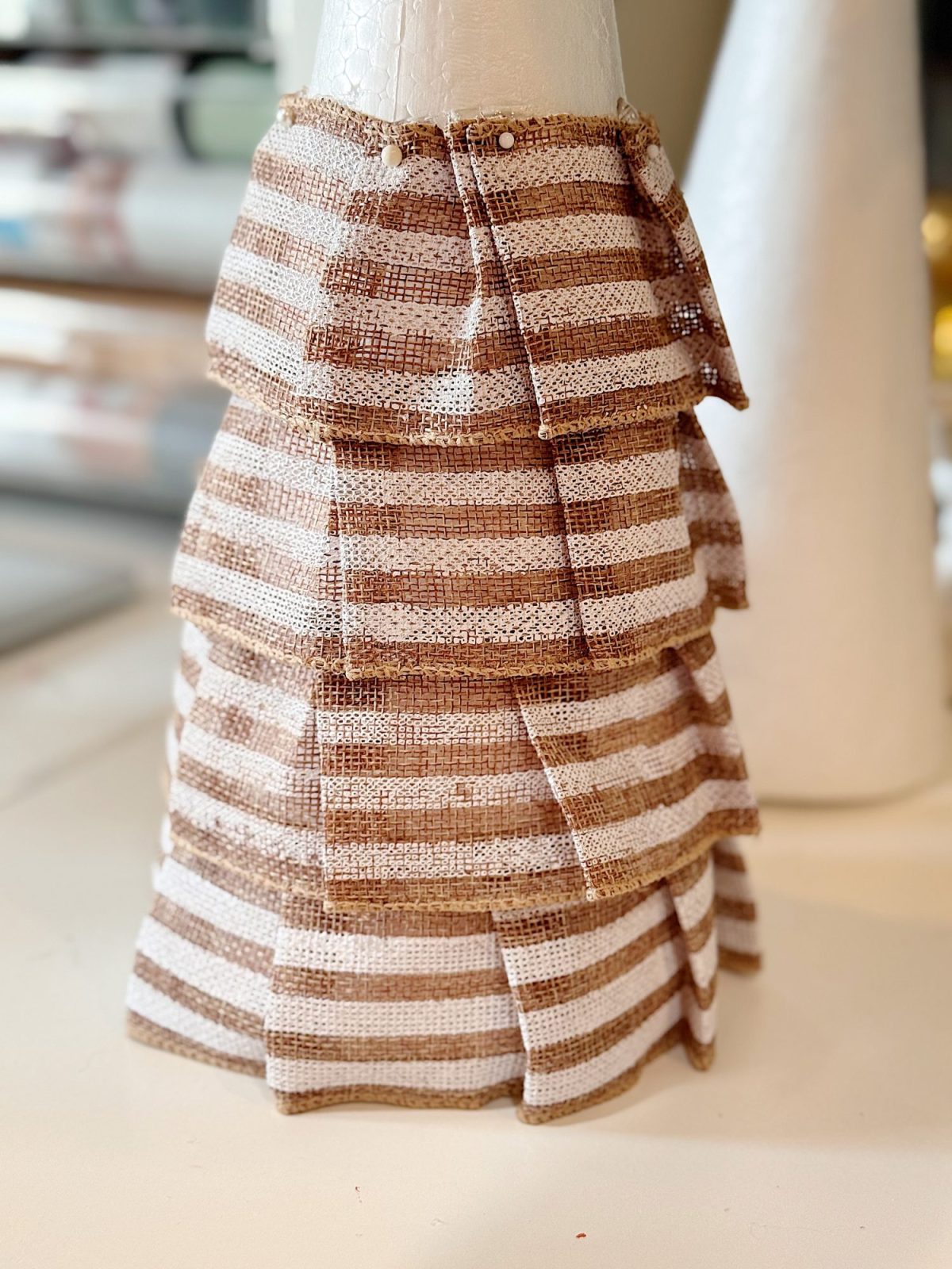 A cone-shaped object covered with layered, brown and white striped burlap fabric strips, resembling a decorative craft project.