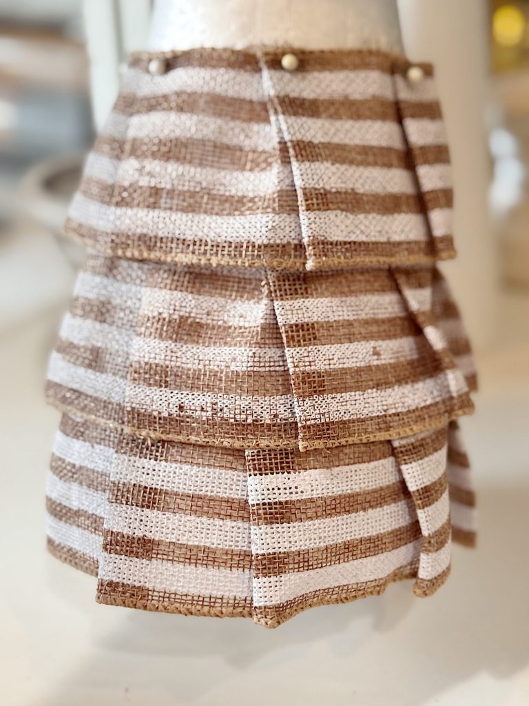 A close-up of layered brown and white striped burlap fabric pinned to a mannequin, arranged in a tiered, ruffled pattern.