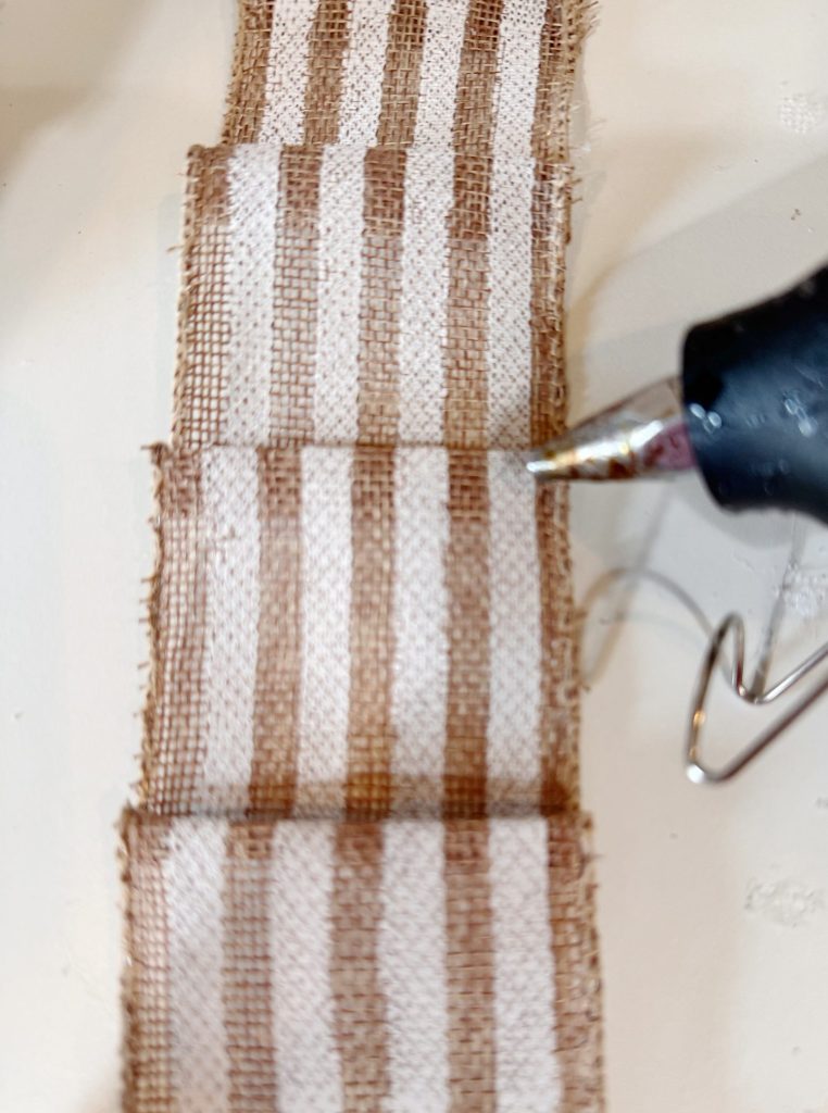 A striped burlap ribbon is being glued in folded sections with a hot glue gun.