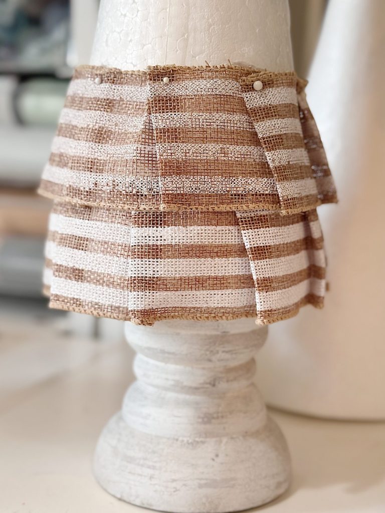 A cone-shaped foam base is covered with two layers of brown and white striped burlap fabric, pinned in place, and displayed on a white wooden stand.