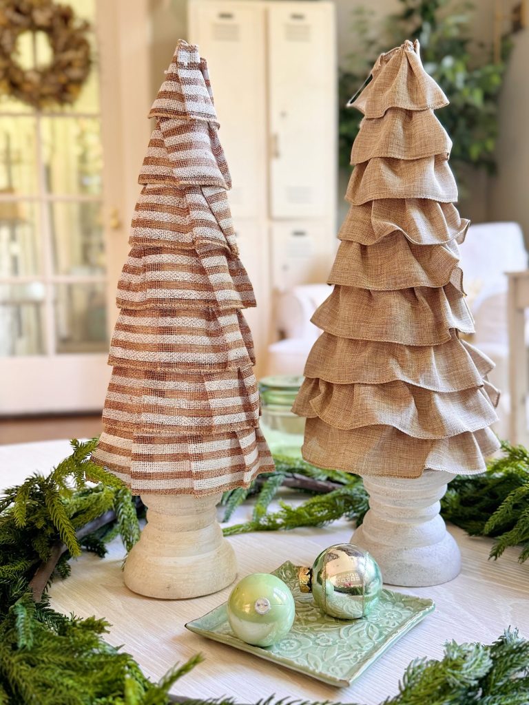 Two fabric Christmas trees with ruffled layers stand on a table, surrounded by greenery, with two decorative ornaments on a square plate in the foreground.