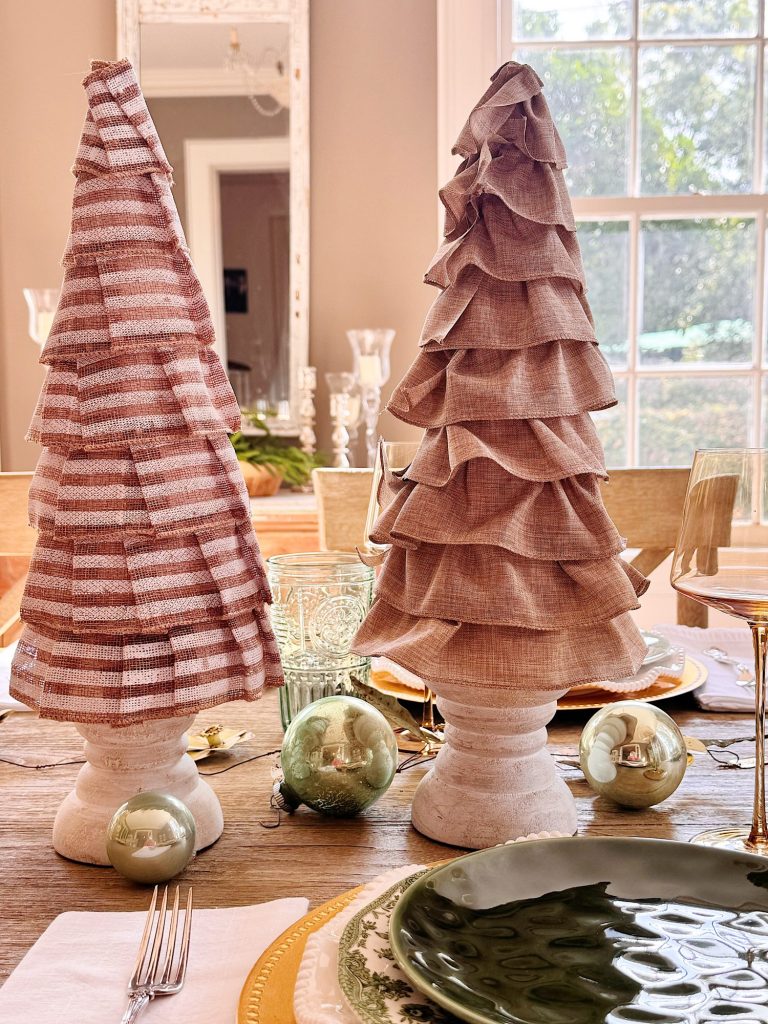 Two decorative fabric Christmas trees sit on a wooden dining table set with plates, glassware, silverware, and green ornaments. A window and mirror are visible in the background.