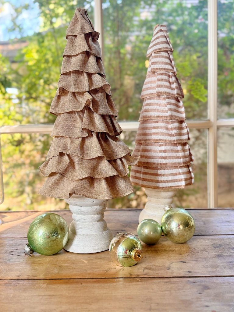 Two decorative fabric Christmas trees and several green ornaments are arranged on a wooden table in front of a window.