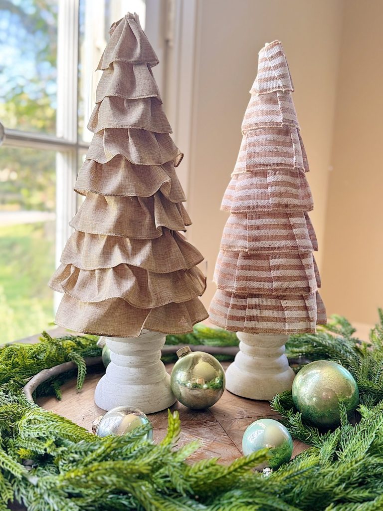 Two fabric Christmas tree decorations on white stands, surrounded by green garland and metallic ornaments, placed on a wooden tray near a window.