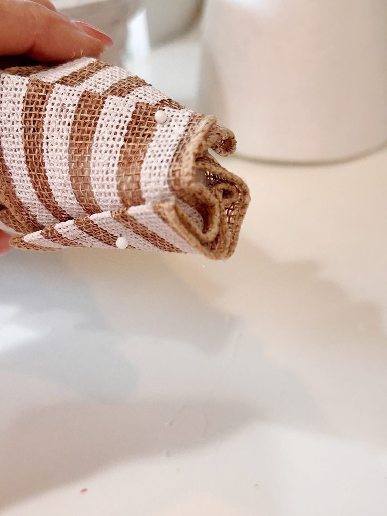 A hand holds a piece of striped burlap fabric rolled up and secured with sewing pins, against a white background.