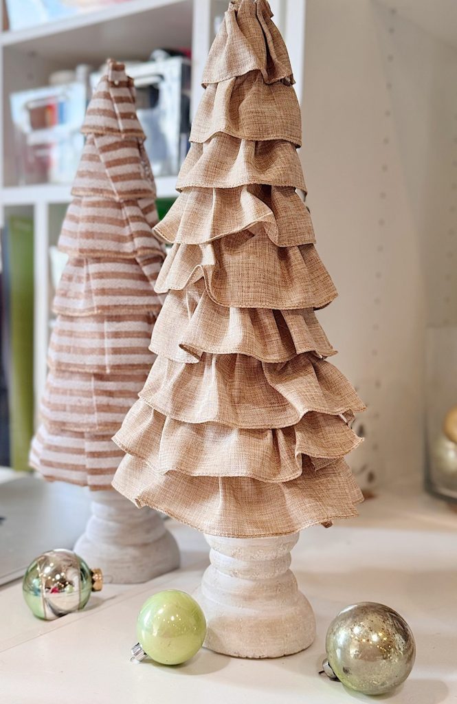 Two fabric Christmas trees with ruffled layers stand on a white surface, accompanied by green and gold ornaments. Shelves with storage containers are visible in the background.