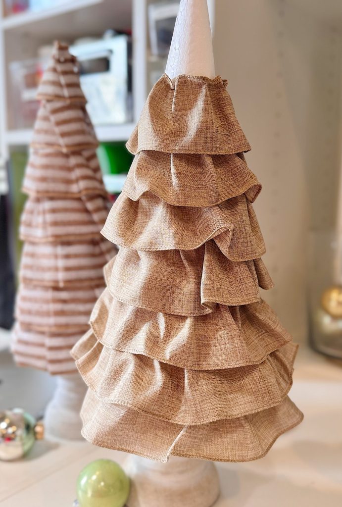 Fabric ruffled tree decoration with beige layered ruffles on a white base, with another similar tree and ornaments in the background.