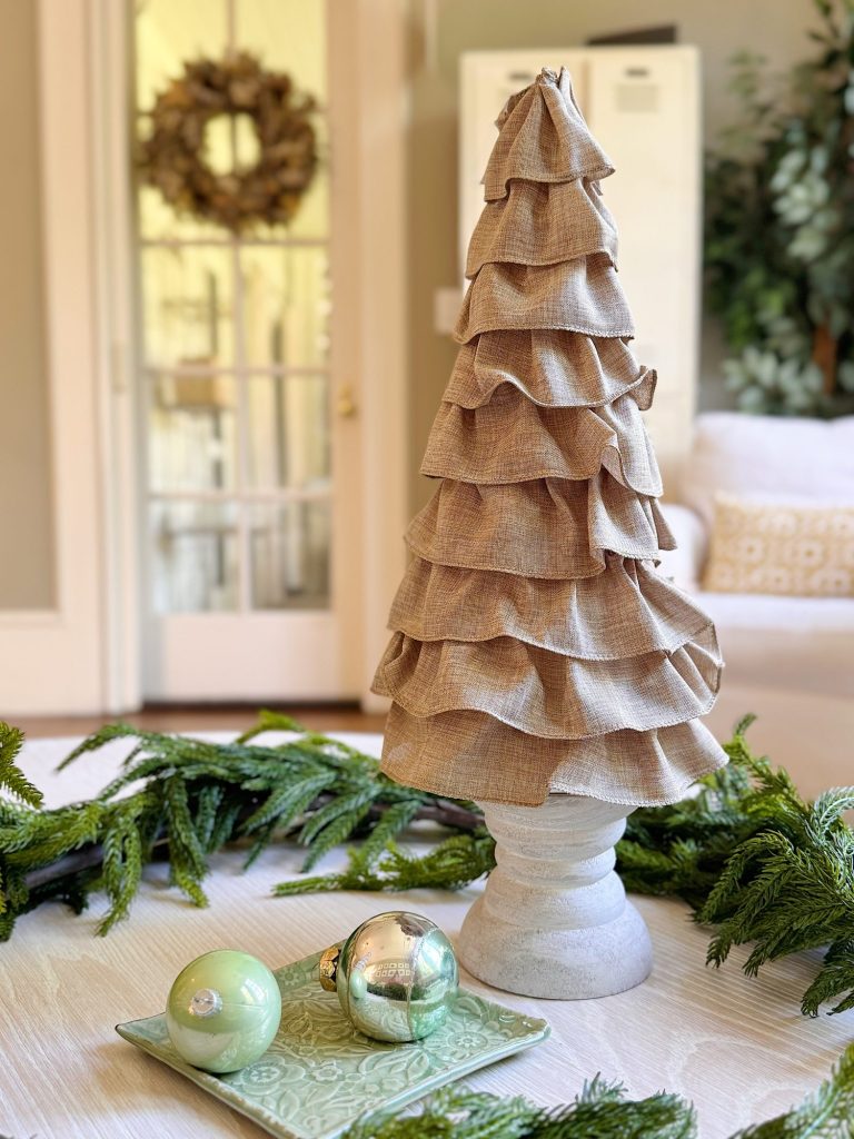 A fabric ruffled cone-shaped decoration sits on a table next to two ornaments on a small tray, with greenery and a living room in the background.
