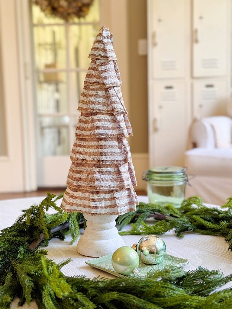 A fabric-covered cone shaped like a Christmas tree sits on a table with a green wreath and ornaments in a bright room.