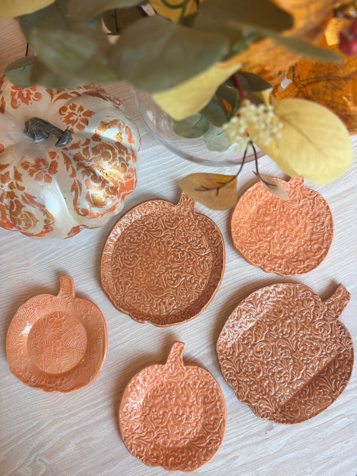 Five orange pumpkin-shaped plates with embossed patterns are arranged on a table next to a decorative white pumpkin and autumn leaves.