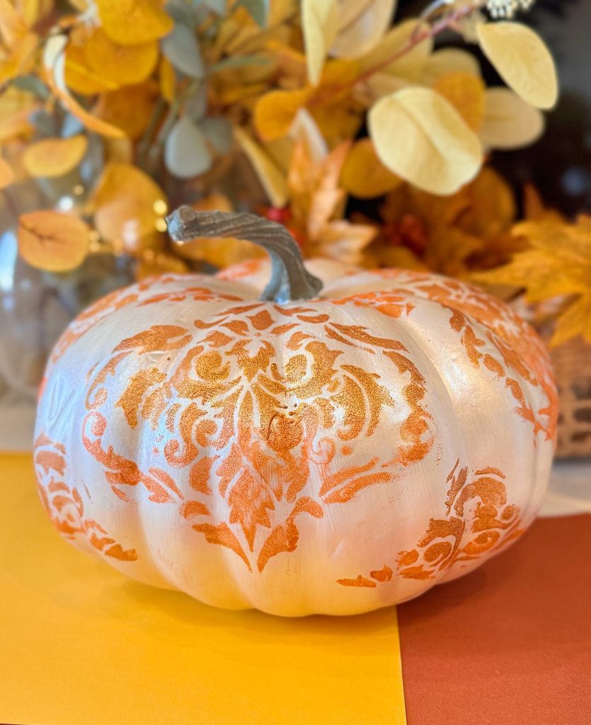 A white pumpkin decorated with an ornate orange floral pattern sits on a surface, with autumn leaves and a vase in the background.