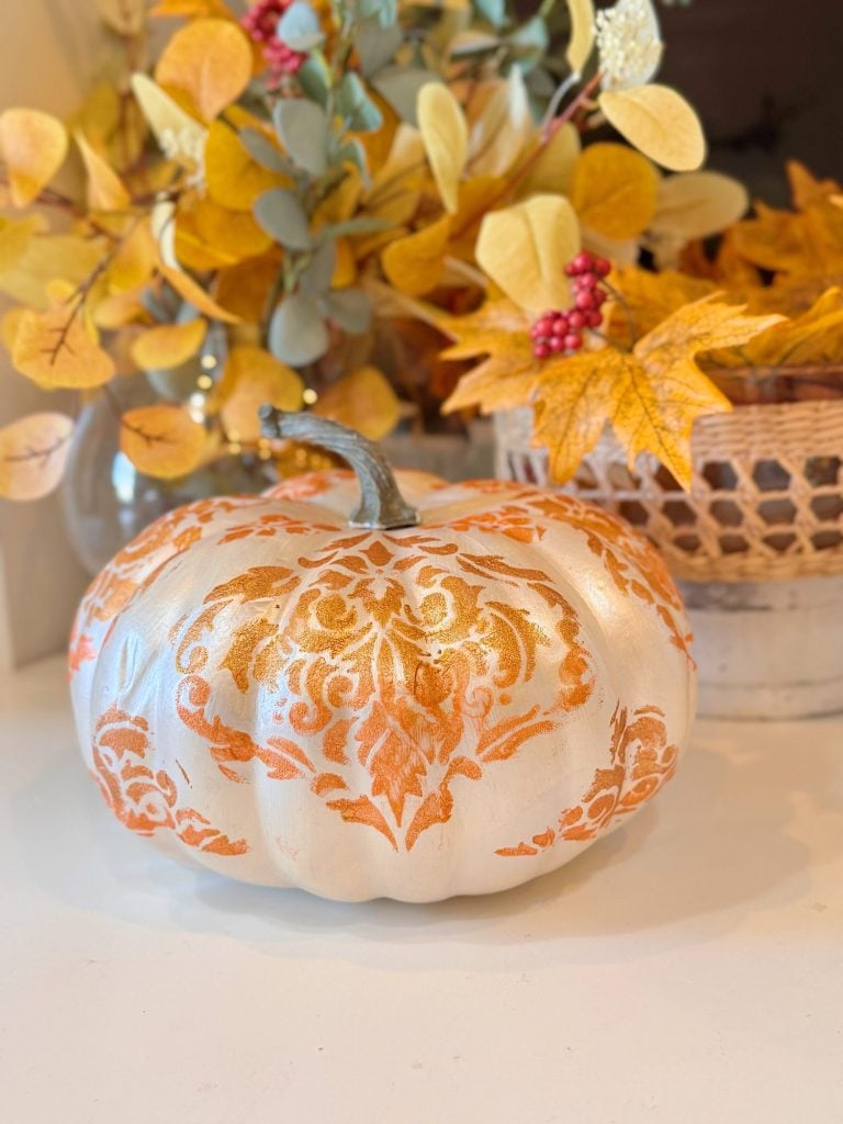 A white pumpkin with an orange damask pattern sits on a white surface, with a basket of autumn leaves and berries in the background.
