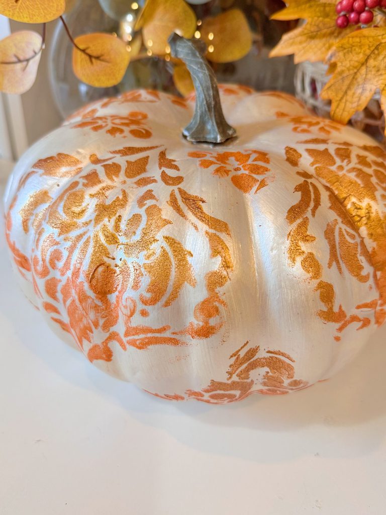 A white pumpkin decorated with an orange ornate stencil pattern, surrounded by artificial autumn leaves.