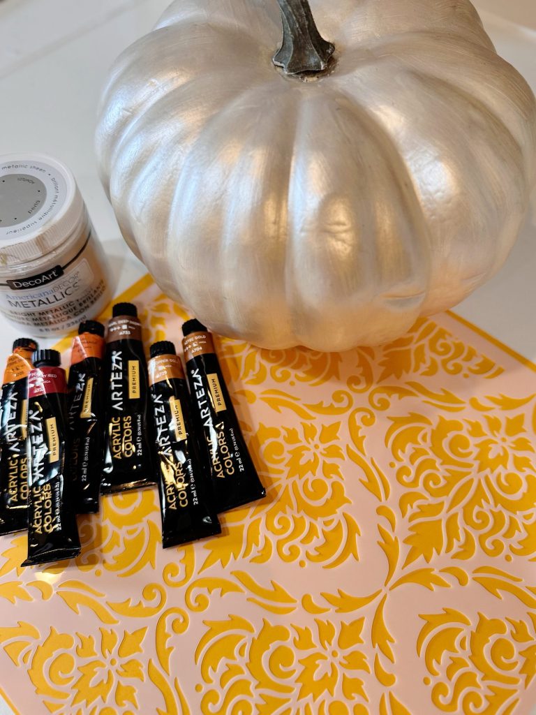 A silver-painted pumpkin, several tubes of acrylic paint, a jar of metallic paint, and a yellow damask stencil are arranged on a flat surface.