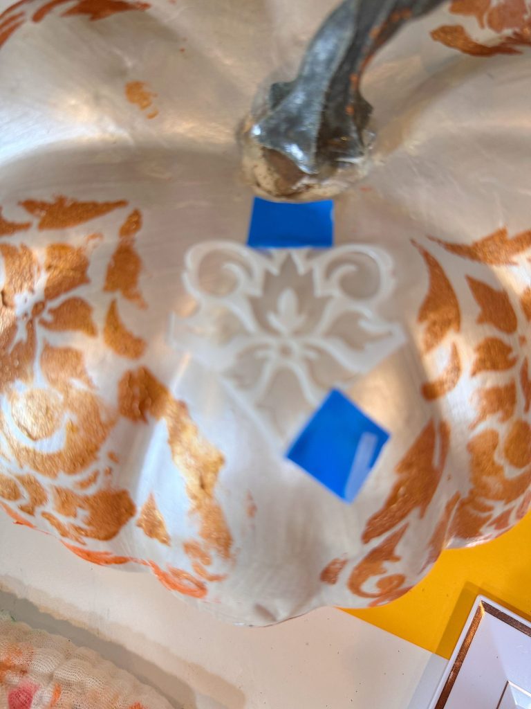 A close-up of a white pumpkin with orange patterns, featuring a decorative white applique taped on with two pieces of blue tape near the stem.