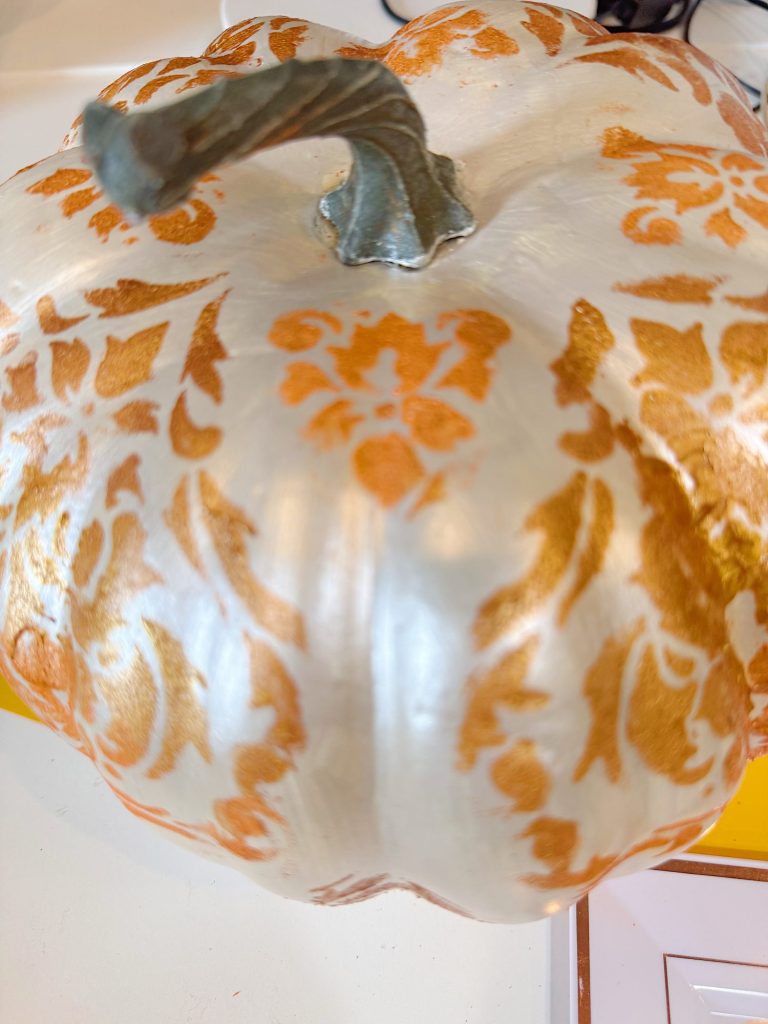 A pumpkin painted white with a gold and orange decorative floral pattern, shown on a white surface.