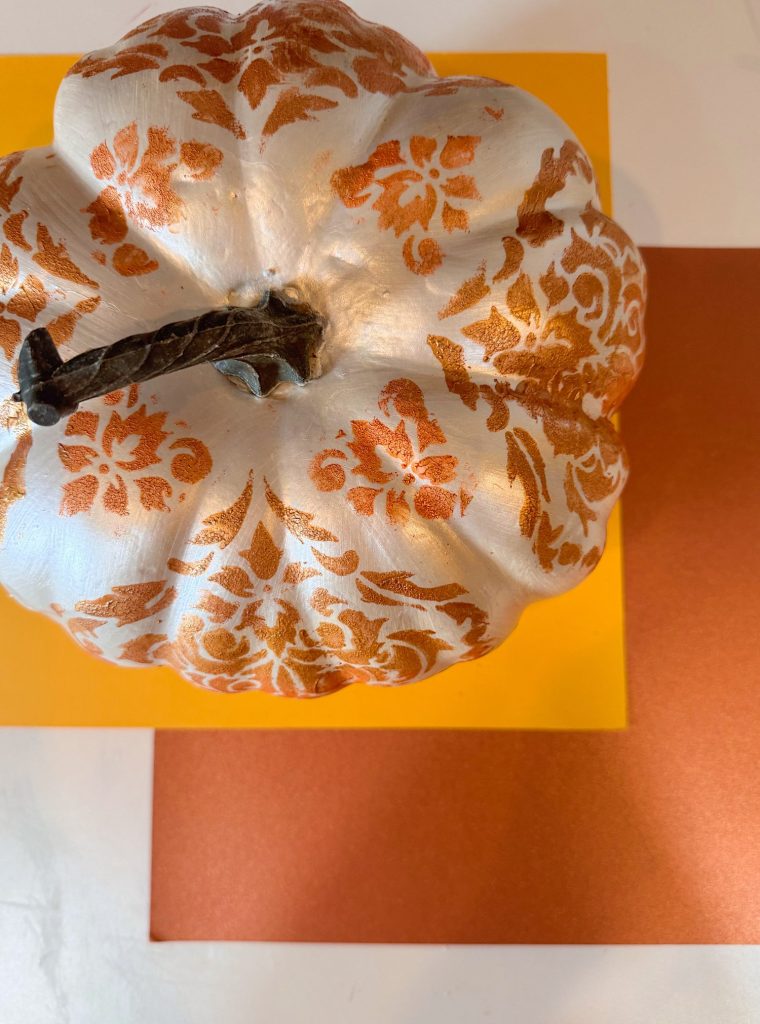 A white pumpkin with orange decorative patterns sits on top of stacked orange and yellow square papers.