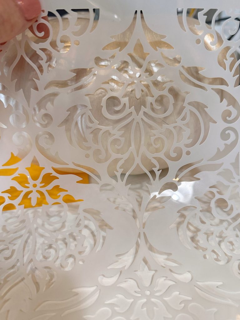A close-up of a hand holding a translucent sheet with an intricate floral and ornamental cut-out pattern, showing a yellow background through part of the design.