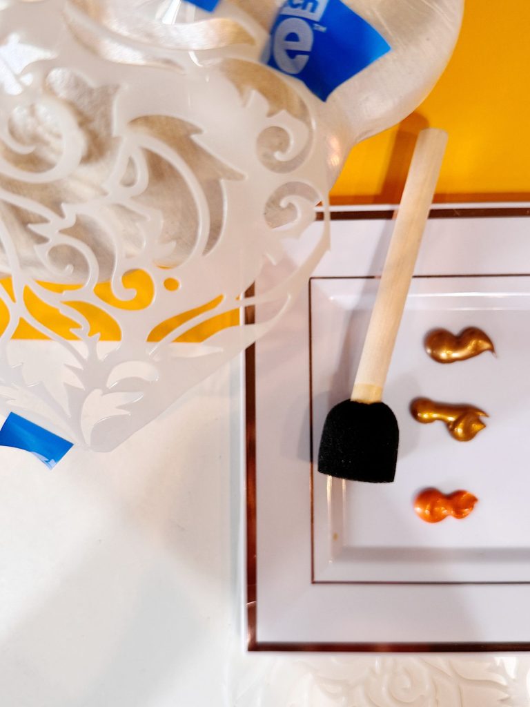A foam brush and a stencil are next to a plate with three metallic paints in gold, bronze, and copper shades.
