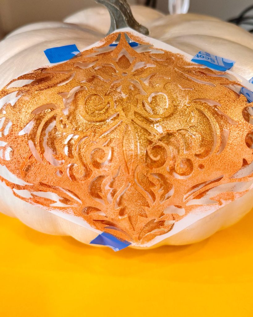 A white pumpkin with a gold ornate stencil pattern being painted on, held in place with blue painter’s tape.