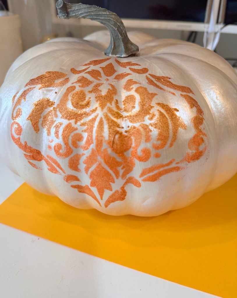 A white pumpkin with an orange intricate damask pattern painted on its surface, displayed on a white and yellow background.