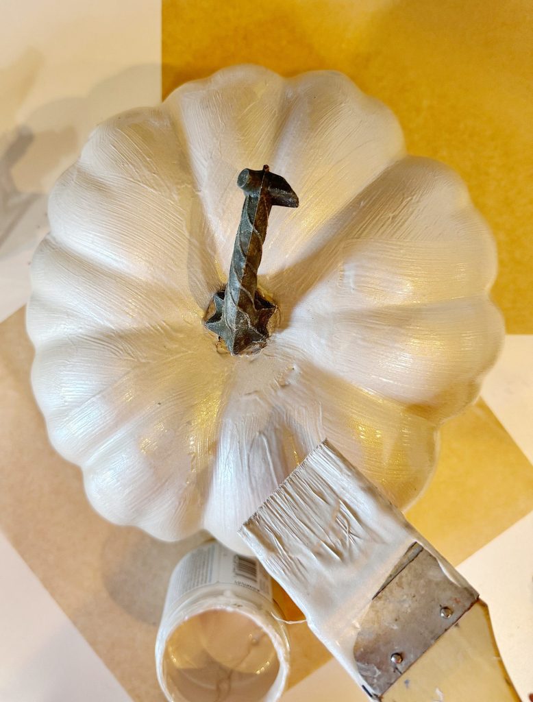 A white pumpkin is being painted with a brush; a small jar of paint is nearby on a yellow and white surface.