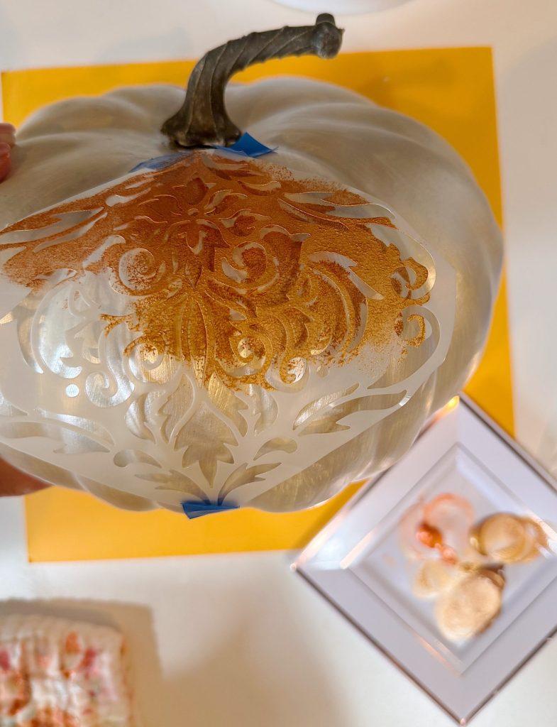 A white pumpkin is being stenciled with a gold ornate pattern. Paint supplies and a plate with paint dabs are nearby on a yellow surface.