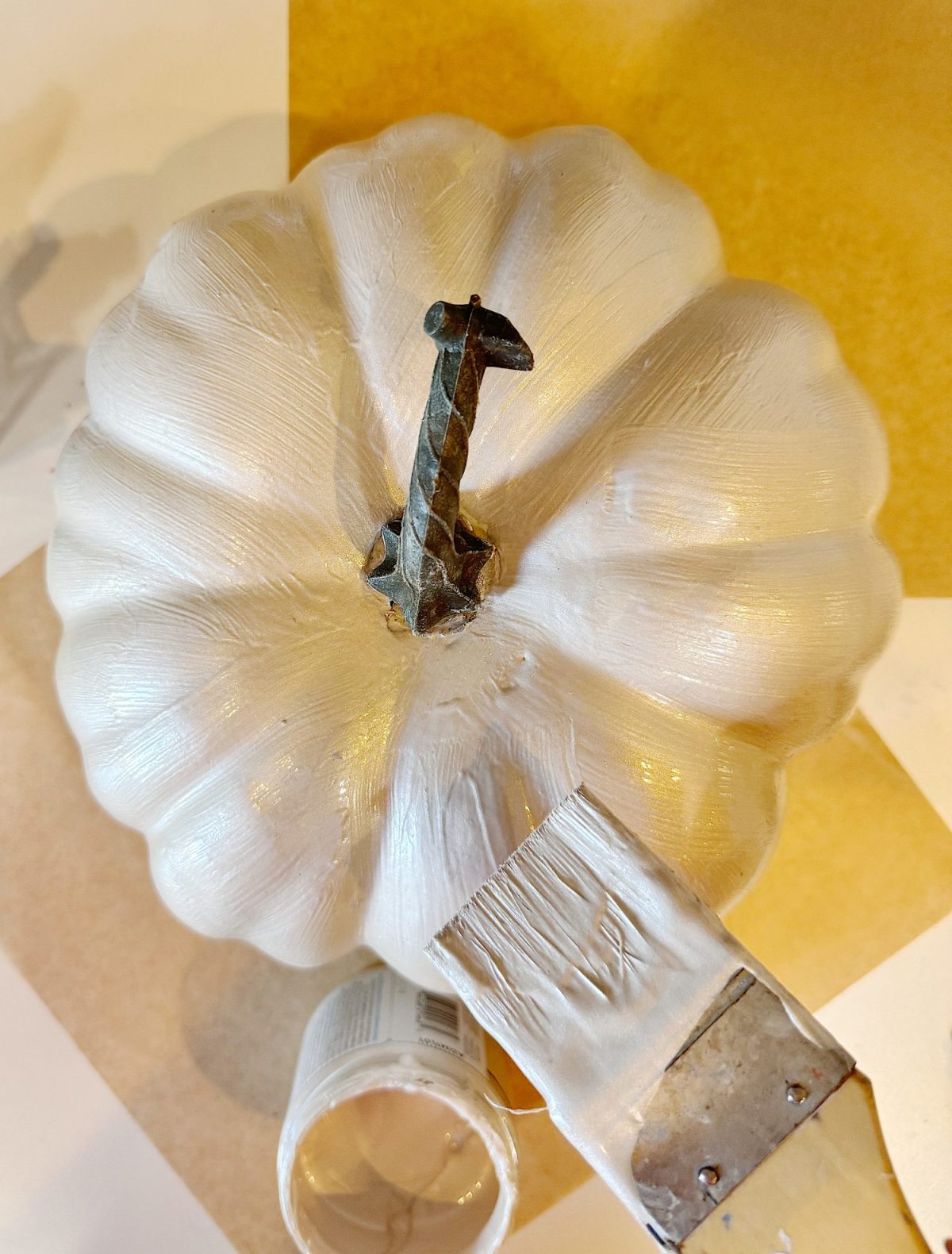 A white pumpkin is being painted with a brush; a small jar of paint is nearby on a yellow and white surface.