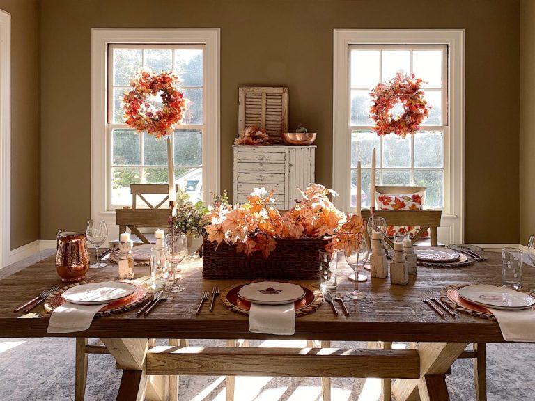 A dining table set for a meal with autumn-themed decorations, including orange leaves, surrounded by wooden chairs and windows adorned with fall wreaths.