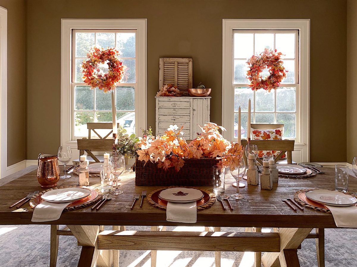 A dining table set for a meal with autumn-themed decorations, including orange leaves, surrounded by wooden chairs and windows adorned with fall wreaths.