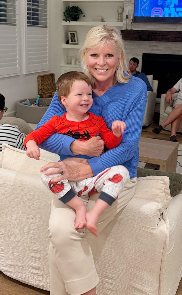 A woman in a blue sweater sits on a couch, smiling and holding a young boy dressed in Spider-Man pajamas, enjoying the holiday season. Other people are in the background.