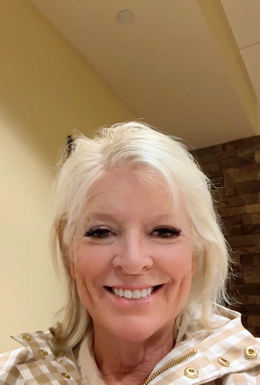 A woman with light hair and a checked jacket smiles at the camera indoors, with beige walls and a stone accent in the background.
