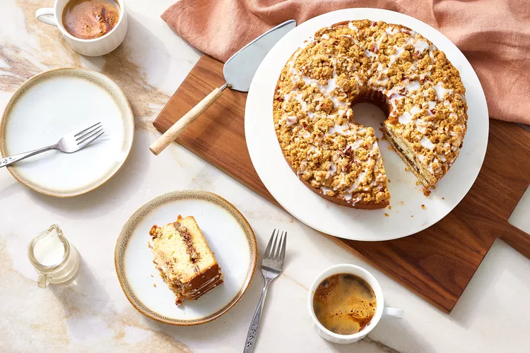 A sliced coffee cake with crumb topping and icing sits on a wooden board, with a serving spatula, two cups of coffee, a small pitcher, and plated cake on a marble surface.