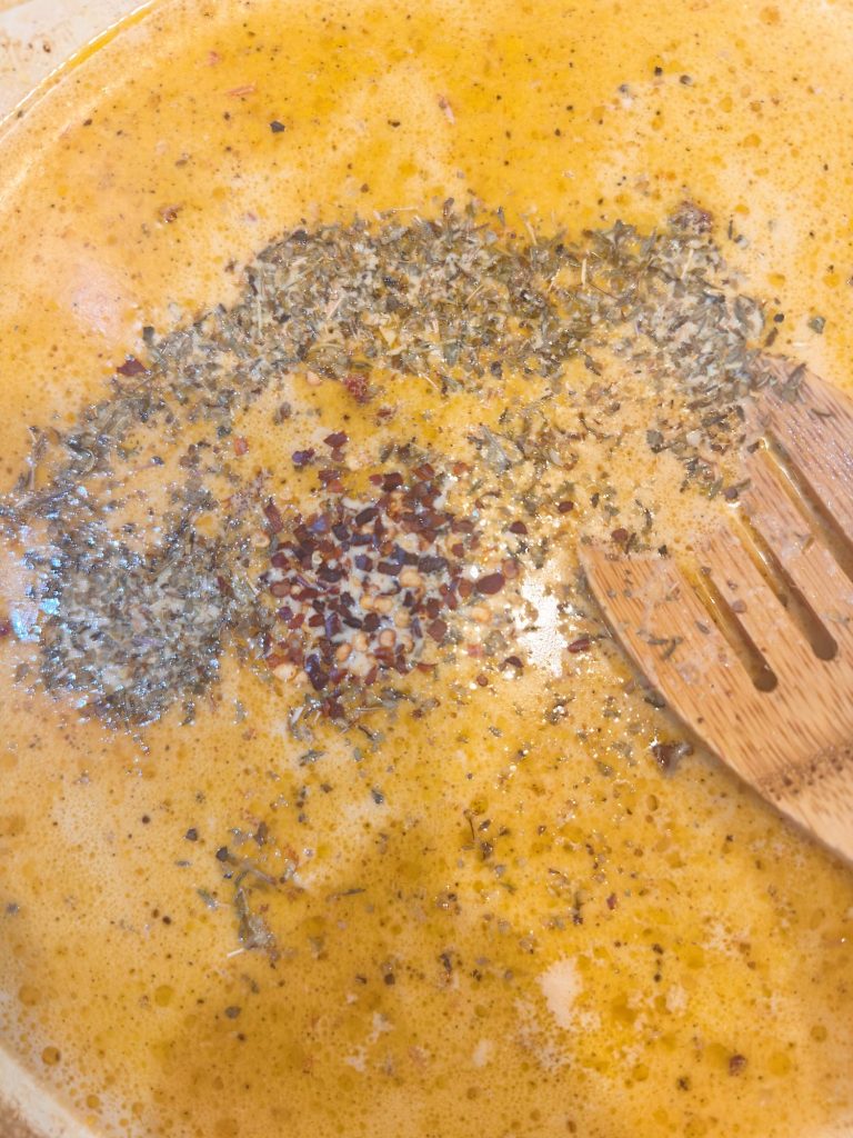 A close-up of a creamy soup being stirred with a wooden spoon, showing dried herbs and red pepper flakes on the surface.