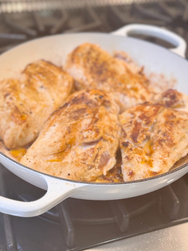 Four browned chicken breasts cooking in a white skillet on a stovetop.