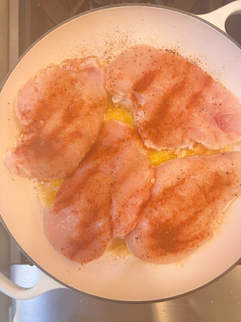 Four raw chicken breasts seasoned with paprika and pepper are cooking in a white skillet with oil on a stovetop.