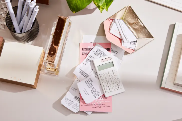 A desk with a stack of receipts, a white calculator, a gold stapler, pens, a small gold envelope, a notepad, and a green plant.