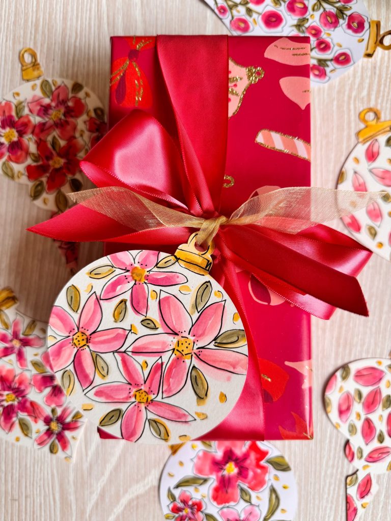 A gift wrapped in red floral paper with a large red ribbon and bow, decorated with a round ornament featuring pink flowers; similar ornaments surround the gift.