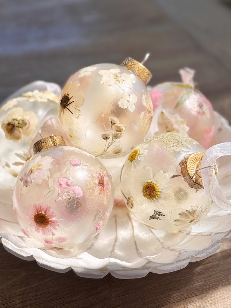 Clear glass ornaments with dried flowers inside are arranged on a scalloped white dish, illuminated by soft natural light.