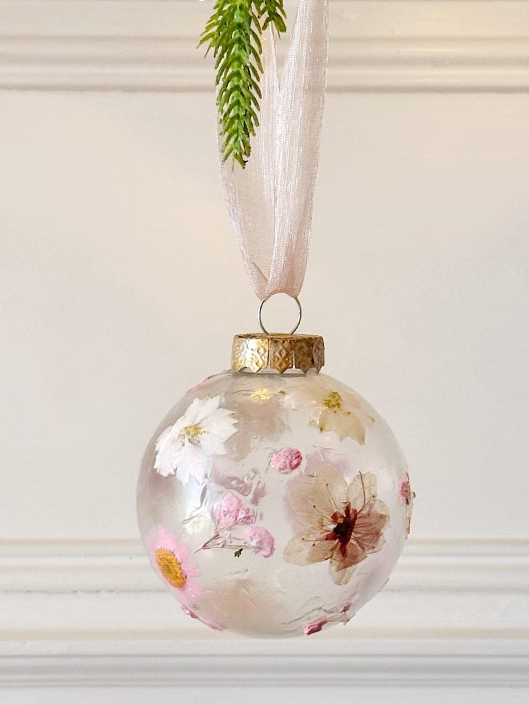 Clear glass ornament with dried flowers inside, hanging by a sheer pink ribbon against a white background with a green fern above.