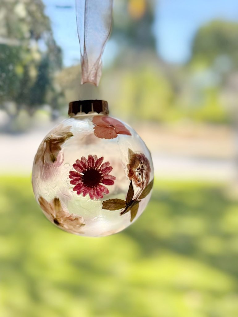 A clear ornament with pressed flowers and leaves inside, hanging by a ribbon in front of a blurred outdoor background.