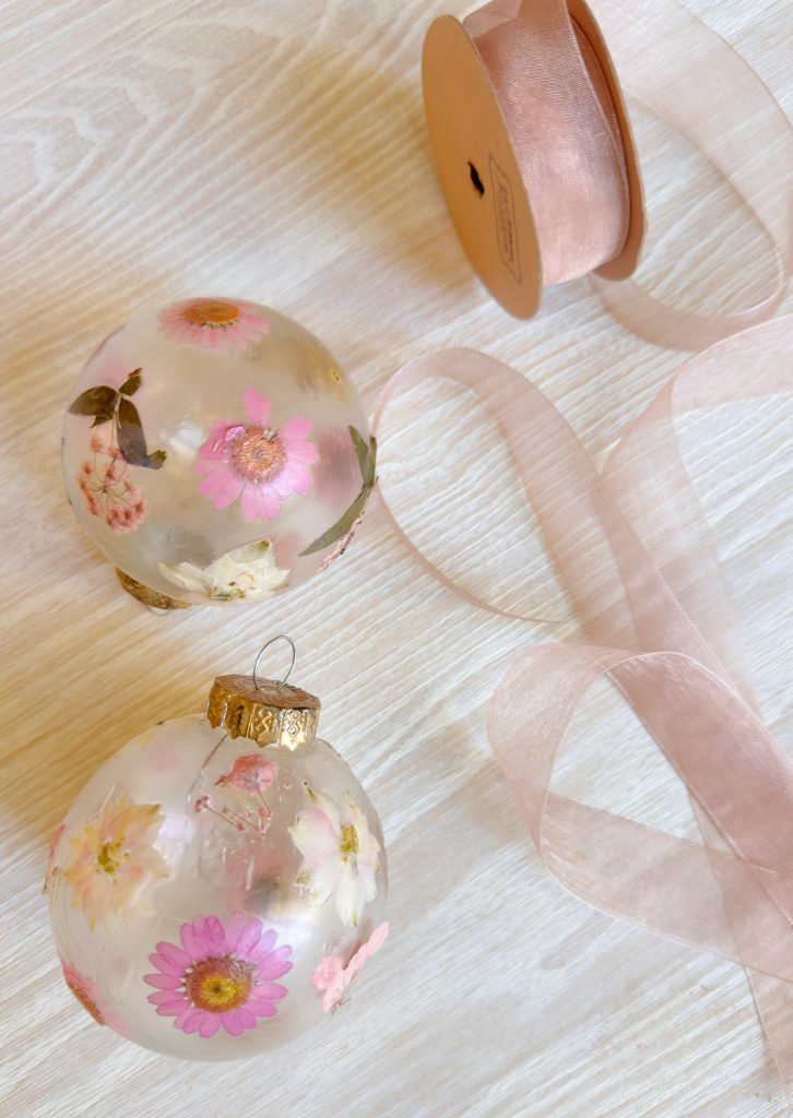 Two clear ornaments filled with pressed flowers rest on a light wood surface next to a spool of pink sheer ribbon and a matching loose ribbon.