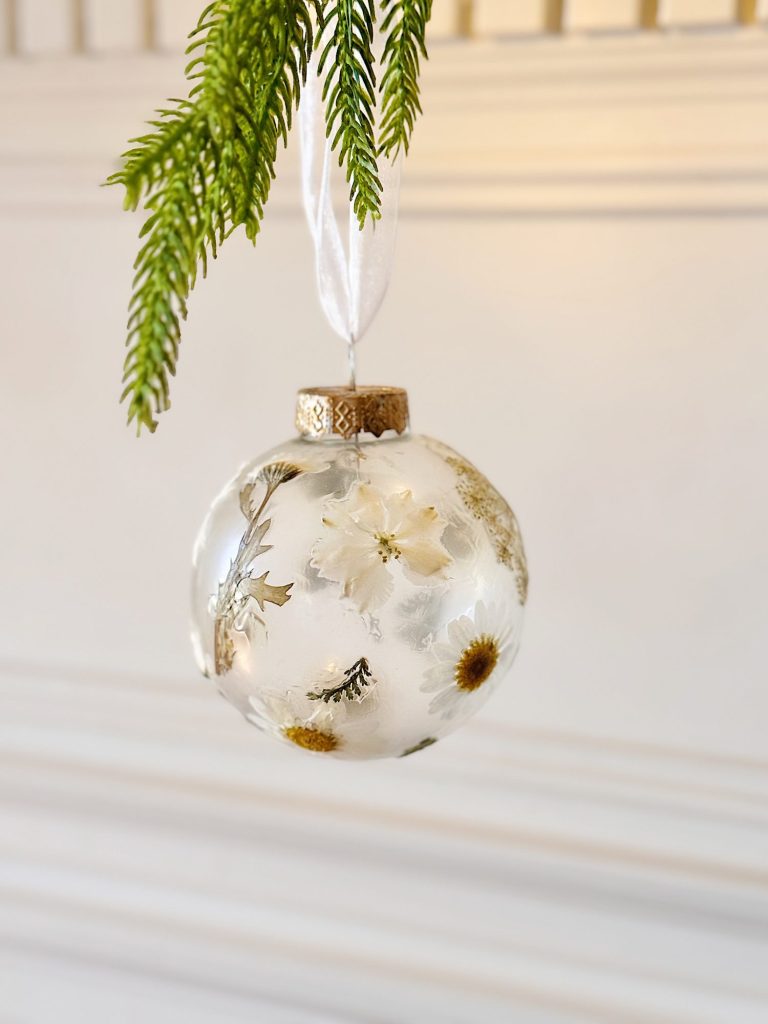 A clear glass ornament with pressed white and yellow flowers inside hangs from a white ribbon, with green pine needles above it.
