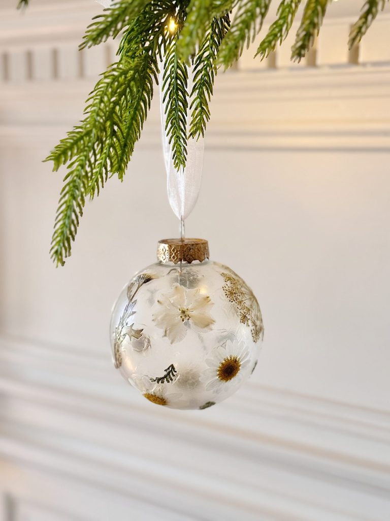 A clear glass ornament with dried flowers inside hangs from a white ribbon on a green pine branch against a white background.