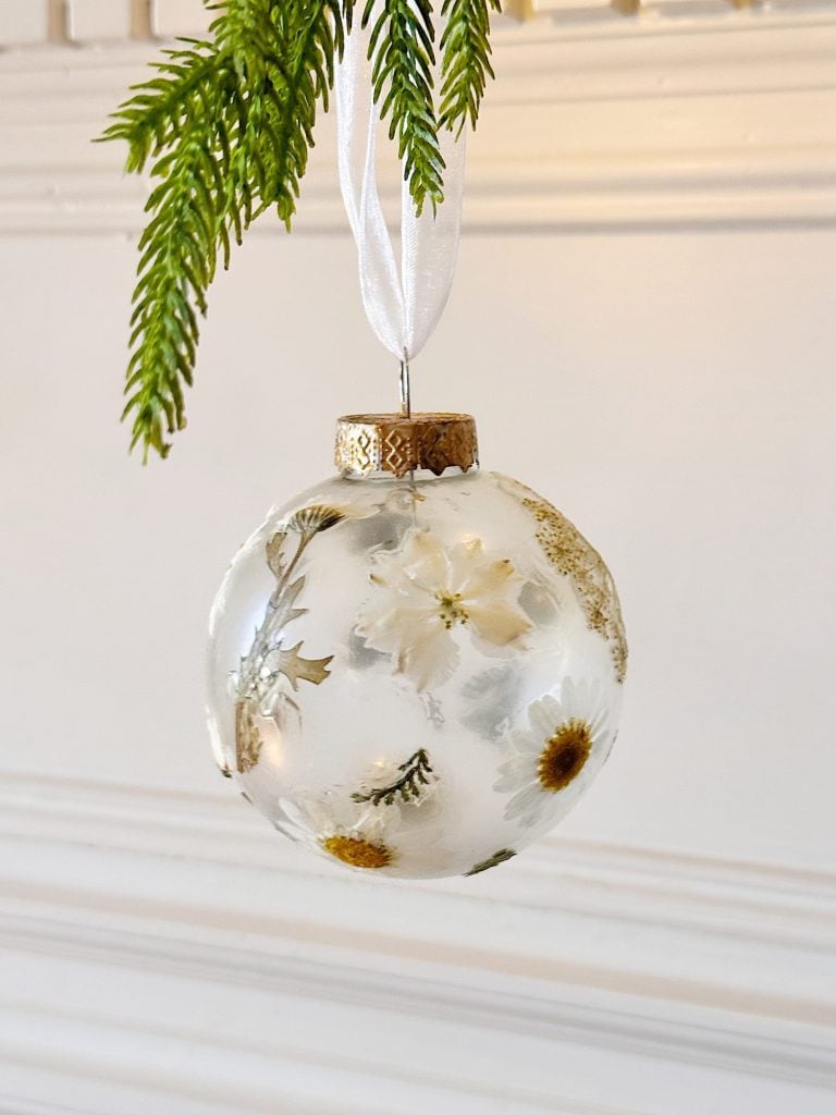 A clear round ornament filled with dried white and yellow flowers hangs from a white ribbon, with green pine needles above.