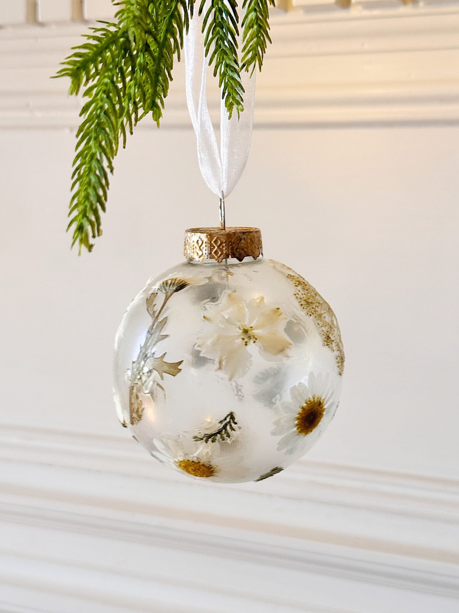 A clear round ornament filled with dried white and yellow flowers hangs from a white ribbon, with green pine needles above.