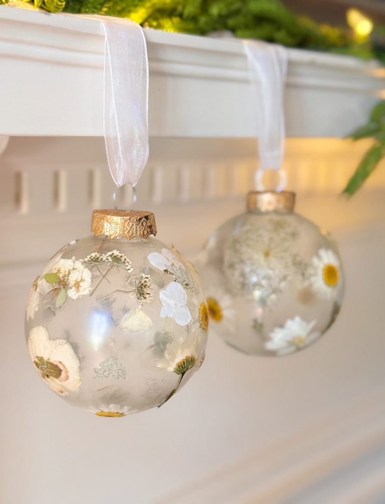 Two clear glass ornaments filled with dried flowers and hung by white ribbon are displayed against a white mantel.