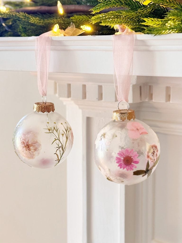 Two clear glass ornaments with dried flowers inside hang by pale pink ribbons from a white mantel, with greenery and string lights above.